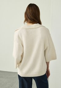 Pull crème avec un col côtelé et des manches courtes, présentant une texture douce et une coupe décontractée, associé à un jean bleu foncé.