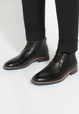 Bottines en cuir noir avec lacets, semelles contrastées marron et bleues, portées avec un pantalon noir retroussé sur un fond clair.
