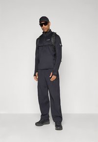 Chaqueta negra para exteriores con cremallera parcial, combinada con pantalones cargo negros de corte holgado. Incluye una mochila táctica y gafas de sol.