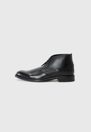 Bottine en cuir noir avec une texture lisse, bout pointu, design à lacets avec deux œillets, et un petit talon empilé. Coutures sur les côtés.