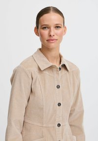 Beige kordjacka med krage, knappfäste fram och två fickor. Har vertikala linjer och en texturerad yta.