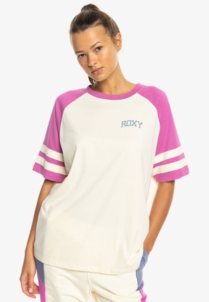 Mladá žena v bežnom krémovom tričku s ružovými rukávmi a logom "ROXY", pózujúca s jednou rukou v vrecku na bielom pozadí.