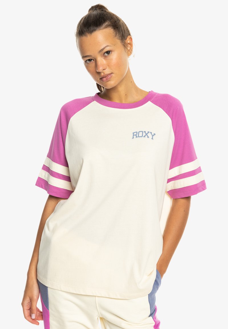 Młoda kobieta w casualowej kremowej koszulce z różowymi rękawami i logo "ROXY", pozująca z jedną ręką w kieszeni na białym tle.