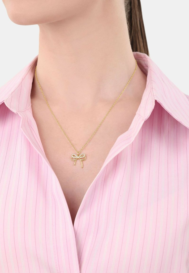 Collar de oro con un colgante en forma de lazo adornado con pequeñas piedras preciosas, llevado sobre una camisa de rayas rosa claro, destacando los detalles del diseño.