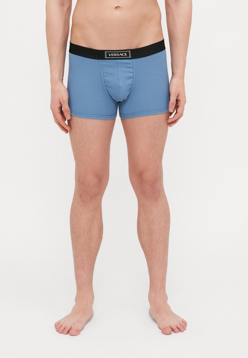 Boxerii scurți pentru bărbați, de culoare albastru deschis, cu un brâu negru al logo-ului Versace, având un material texturat și un stil ajustat.