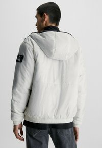 Doudoune blanche avec une capuche, une taille élastique et des accents noirs. Présente un patch logo CK sur la manche gauche. Tissu texturé.