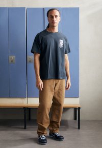 Svart bomull t-skjorte med grafikk på forsiden og korte ermer, sammen med brune bukser med synlige sømmer og hardware-detaljer. Svart joggesko.
