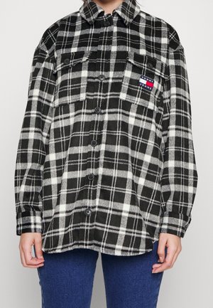 Osoba nosząca czarną i białą koszulę w kratę zapinaną na guziki z kieszeniami na piersi i logo Tommy Jeans, w zestawieniu z niebieskimi dżinsami.