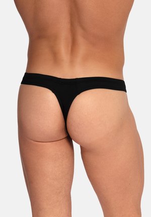 Männlicher Oberkörper und obere Beine von hinten, trägt schwarze Tanga-Unterwäsche vor einem schlichten hellen Hintergrund.