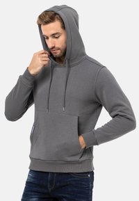 Grauer Hoodie mit einer Fronttasche, verstellbarem Kordelzug, gerippten Bündchen und einer weichen Textur. Das Modell trägt ihn mit dunklen Jeans.