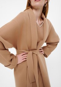 Beige-vaatepeite, joka on valmistettu pehmeästä kankaasta, avoimella edustalla, leveillä hihoilla ja solmitulla vyötärövyöllä. Siinä on hienovarainen tekstuuri.