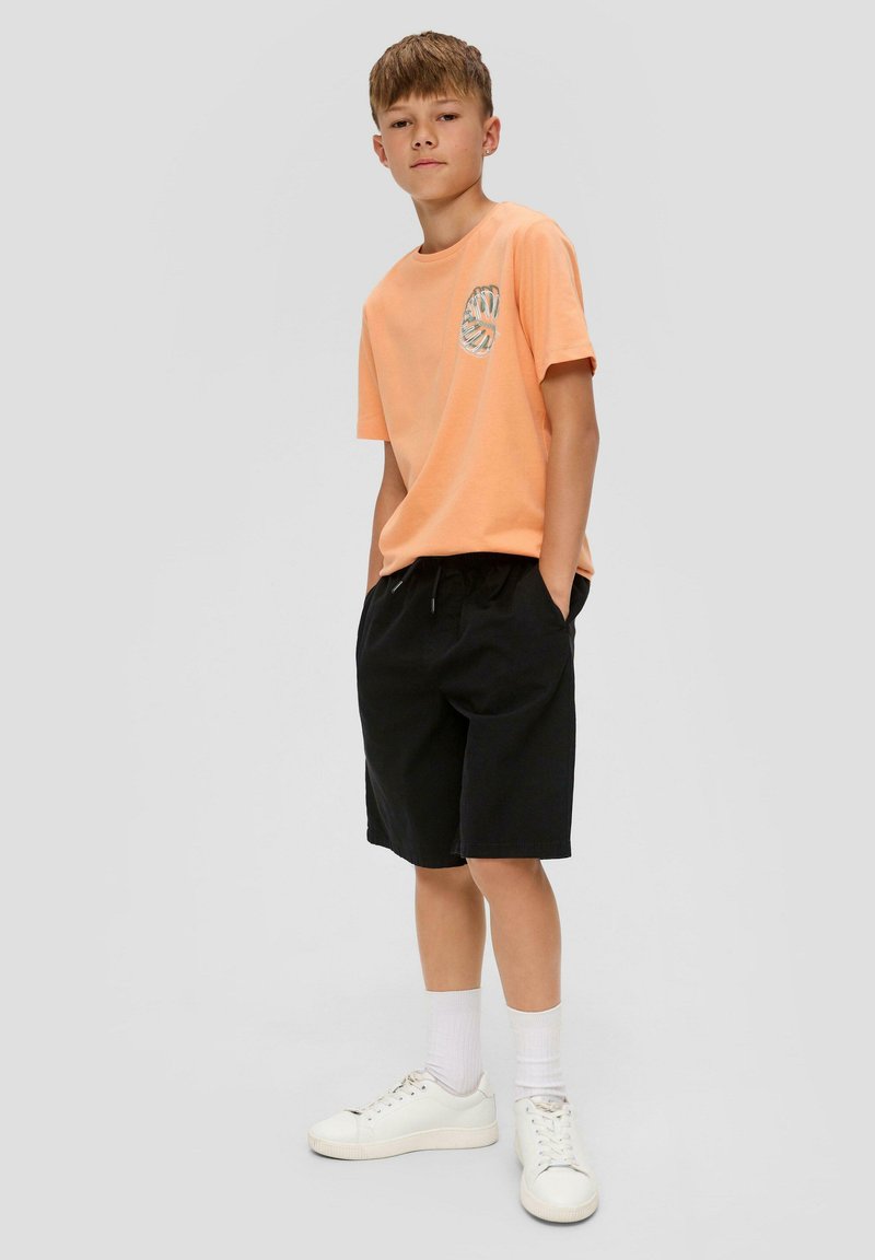 Een oranje katoenen t-shirt met een grafisch ontwerp aan de voorkant, gecombineerd met zwarte shorts en witte sneakers, staand op een grijze achtergrond.