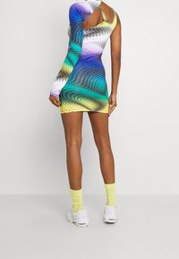 Kobieta ubrana w wielokolorową sukienkę bodycon z jednym rękawem, w żółtych skarpetkach i białych tenisówkach, stojąca na tle jasnego, jednolitego tła.