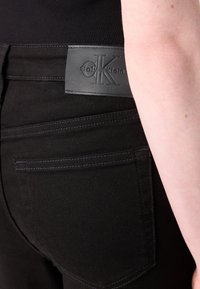 Blugi negri din denim cu buzunar în spate și patch cu logo-ul Calvin Klein pe talie, purtați de o persoană cu piele deschisă la culoare vizibilă pe partea dreaptă.