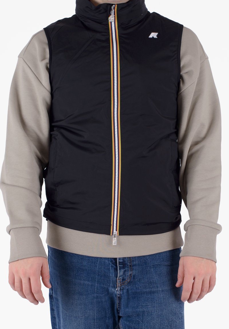 Mann trägt ein beigefarbenes Langarmhemd unter einer schwarzen ärmellosen Weste mit Reißverschluss und mehrfarbigem gestreiftem Reißverschluss sowie blauer Jeans.