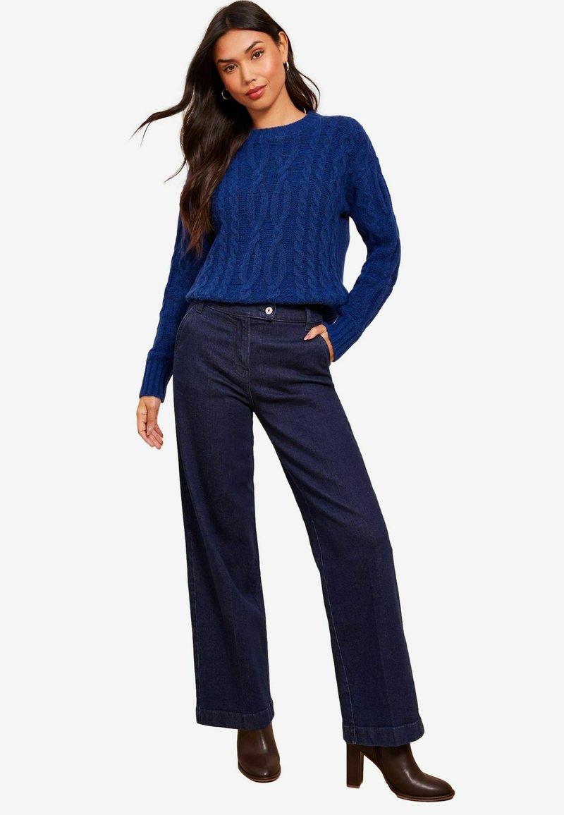 Modra pletena pulover s rebrastimi manšetami, v kombinaciji s širokimi temnimi jeans hlačami. Model nosi rjave škornje s peto, stoji na nevtralni ozadju.