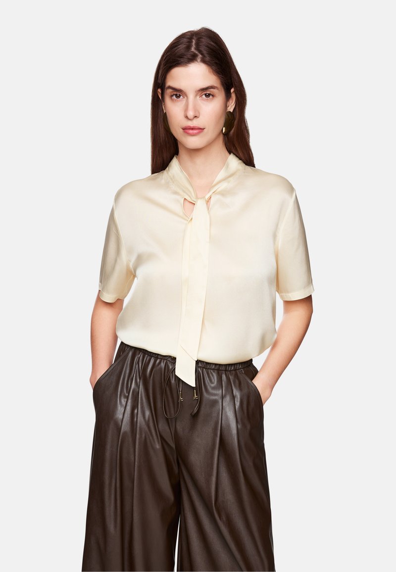 Elfenbeinfarbene Seidenbluse mit einer Krawattenhalsdetail, kurzen Ärmeln und einem subtilen Glanz. Kombiniert mit dunkelbraunen Lederhosen mit einem Kordelzugbund.