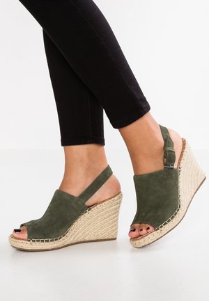 Zielone zamszowe sandały na koturnie z otwartym palcem, paskiem na pięcie i plecionym jucie na koturnie. Czarne spodnie widoczne poniżej.