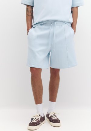 Osoba koja nosi svijetloplave kratke hlače, bijele čarape i tamnosmeđe tenisice, stoji sa rukama u džepovima na jednobojnoj pozadini.