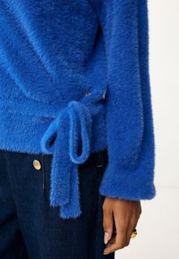 Blå fluffig tröja med knytning i midjan, långa ärmar och ribbade muddar, tillsammans med mörka jeans med guldknappar.