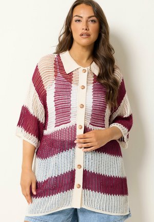 Kvinde iført en løs, knappet, åbenstrikket cardigan med beige krave og skiftende burgunderrøde og hvide striber, kombineret med lyseblå jeans.