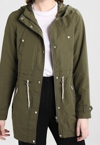 Henkilö, joka on pukeutunut oliivinvihreään huppariin, jossa on nyöritykset, hopeiset vetoketjut ja painonapit, valkoisen paidan ja mustien housujen päälle.