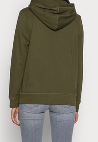 Sudadera con capucha de color verde oliva, con capucha ajustable con cordón, textura suave y puños acanalados, combinada con vaqueros ajustados en gris claro.