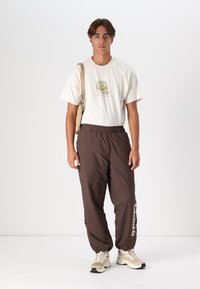 Light beige t-shirt with graphic print, paired with loose-fitting brown nylon pants. Model wears neutral-toned sneakers and a tan shoulder bag.