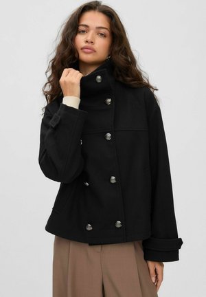 Une femme présente un manteau noir croisé à col montant et boutons argentés, en tenant le col devant un fond de studio uni.
