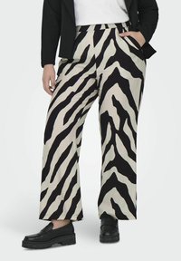Beige wijde broek met een gedurfde zwarte zebra-streep patroon, met zijzakken en een ontspannen pasvorm. G gedragen met zwarte schoenen.
