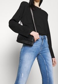 Bolso cruzado de imitación cuero negro con un patrón texturizado de cocodrilo, cadena plateada, combinado con unos jeans desgastados de color azul claro y una blusa negra.