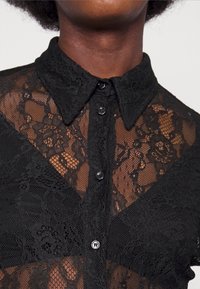 Chemisier en dentelle noire avec un col haut, une fermeture à boutons et des motifs floraux complexes. Le tissu est semi-transparent avec des détails texturés.