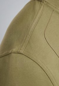 Tessuto di felpa verde oliva con una superficie liscia, caratterizzato da cuciture visibili e un collo a costine. Dettaglio ravvicinato della spalla.