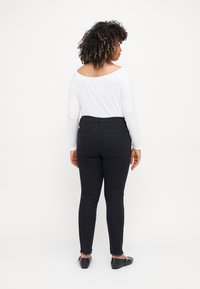 Jeans skinny negros de talle alto, con un dobladillo deshilachado y bolsillos traseros, combinados con una blusa blanca de manga larga. Modelo mostrado desde atrás.