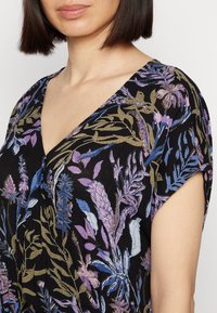 Femme portant une robe noire à encolure en V avec des motifs floraux violets, bleus et beiges et des manches courtes, mettant en valeur le cou et le haut des bras.