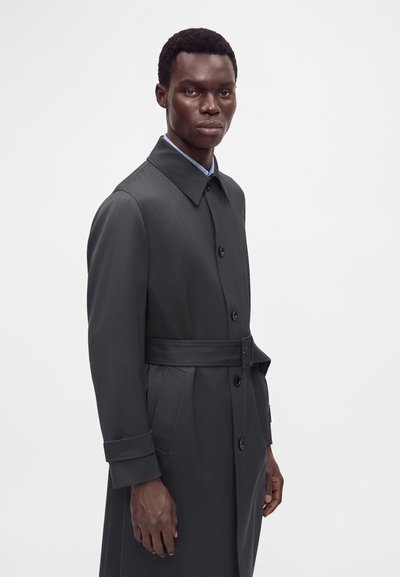 Jeune homme debout portant un trench-coat ceinturé gris foncé avec des boutons et un col de chemise bleu clair visible.