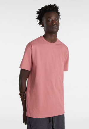 Uomo di colore in piedi con le mani dietro la schiena, indossa una maglietta rosa a maniche corte semplice e pantaloni scuri su uno sfondo bianco.