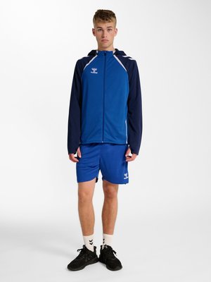 Teenager-Junge trägt eine blaue und marineblaue Sportjacke mit Logo, passende blaue Shorts, weiße Socken und schwarze Sneakers, steht vor weißem Hintergrund.