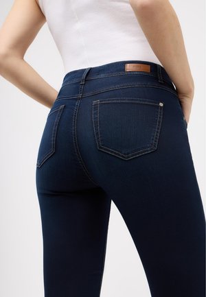 Jean en denim bleu foncé à coupe slim, avec poches arrière et un patch en cuir marron à la ceinture. Les détails de couture visibles améliorent le design.