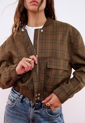 Veste bomber en plaid marron avec une fermeture éclair sur le devant, un col côtelé et une poche. Portée sur une chemise blanche et accompagnée de jeans en denim bleu.