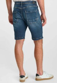 Homme portant un short en denim bleu et des baskets blanches, debout de dos face à l'appareil photo sur un fond clair uni.