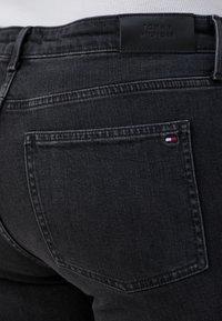 Mørke denimjeans med nærbilde av baklommens design og et merke med lærlabel, samt en liten fargeaksent nær lommens søm.