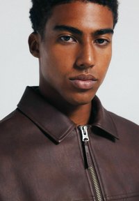 Braune Lederjacke mit Reißverschluss, Kragen und strukturierter Oberfläche. Verfügt über silberne Beschläge und eine strukturierte Passform.