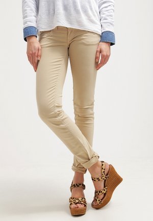 Person trägt beige Slim-Fit-Hosen, an den Knöcheln hochgekrempelt, und Leopardenmuster-Keilsandalen, steht mit gekreuzten Beinen.