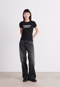 Svart kortermet T-skjorte med "tommy jeans" logo, kombinert med løstsittende mørkegrå denimjeans og svarte sko, på en ensfarget bakgrunn.
