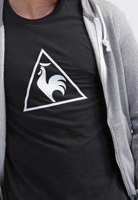 Svart t-shirt med en stor vit triangulär logotyp med en tuppsilhuett i mitten. Materialet ser slätt ut, med en figursydd design.
