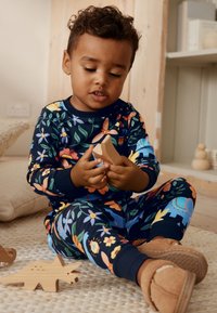 Set pyjamakleding in marineblauw met bloemmotieven, zachte stof en geribbelde manchetten. Kind houdt een houten speelgoeddinosaurus vast op een gestructureerd tapijt.