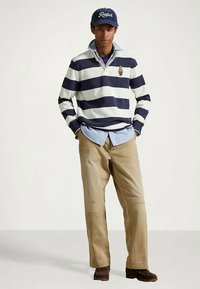 Jeune homme portant un polo rayé bleu marine et blanc, une chemise bleue à boutons, un pantalon beige, des chaussures marron et une casquette "Ralph" bleu marine, se tenant les mains dans les poches.