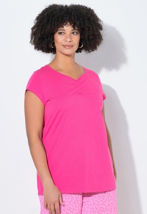 Frau mit lockigem Haar, die goldene Creolen, ein hellrosa Kurzarmtop und hellrosa Leopardenmuster-Hose trägt, steht vor einer weißen Wand.