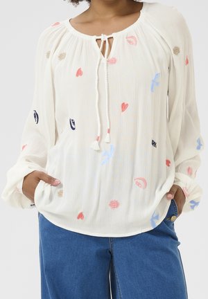 Vrouw draagt een losse witte blouse met kleurrijke geborduurde vormen, lange mouwen en een striksluiting bij de hals, gecombineerd met een blauwe spijkerbroek.